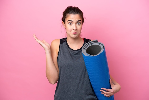 Jeune femme sportive brésilienne allant à des cours de yoga tout en tenant un tapis isolé sur fond rose ayant des doutes tout en levant les mains