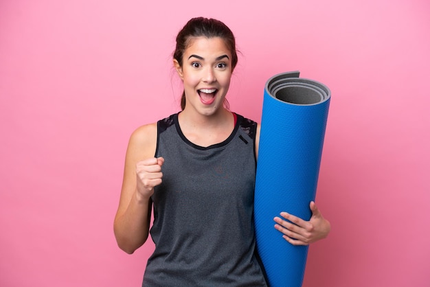 Jeune femme sportive brésilienne allant à des cours de yoga tout en tenant un tapis isolé sur fond rose célébrant une victoire en position de vainqueur