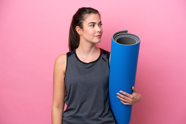 Jeune femme sportive brésilienne allant à des cours de yoga tout en tenant un tapis isolé sur fond rose regardant sur le côté