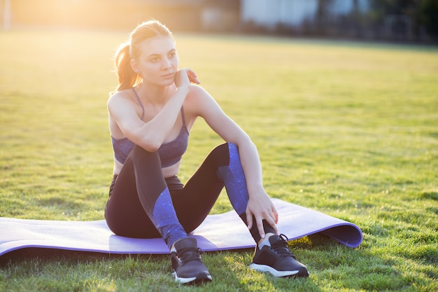 Jeune femme sportive dans des vêtements de sport assis sur un tapis d'entraînement avant de faire des exercices sur le terrain au lever du soleil.