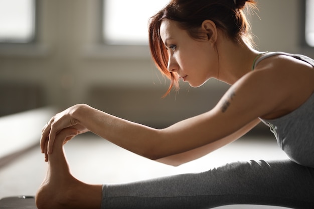 Jeune femme sportive faisant des exercices d'étirement de yoga assis dans une salle de sport près de fenêtres lumineuses.