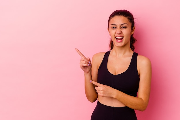 Jeune femme sportive isolée sur un mur rose pointant avec l'index vers un espace de copie, exprimant l'excitation et le désir