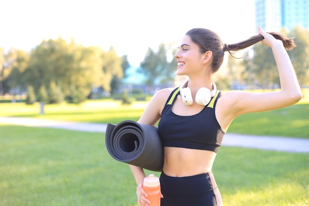 Jeune femme sportive joyeuse marchant dans un parc urbain tenant un tapis de fitness.