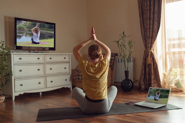 Jeune femme sportive prenant des cours de yoga en ligne et s'entraînant à la maison tout en étant en quarantaine. Concept de mode de vie sain, de bien-être, d'être en sécurité pendant la pandémie de coronavirus, à la recherche d'un nouveau passe-temps.