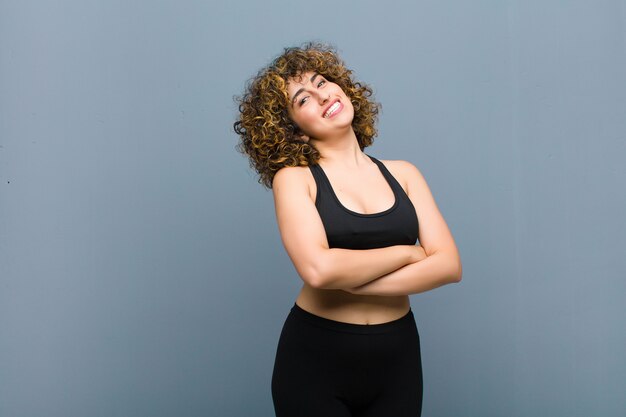 Jeune femme sportive rire joyeusement avec les bras croisés, avec une pose détendue, positive et satisfaite contre le mur gris