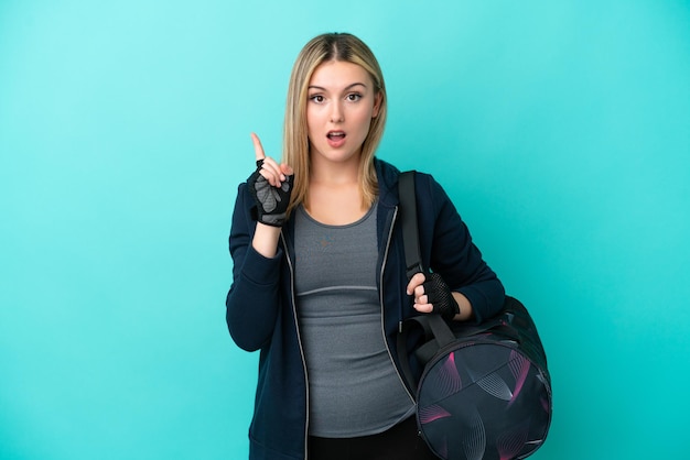 Jeune femme sportive avec sac de sport isolé sur fond bleu pensant une idée pointant le doigt vers le haut