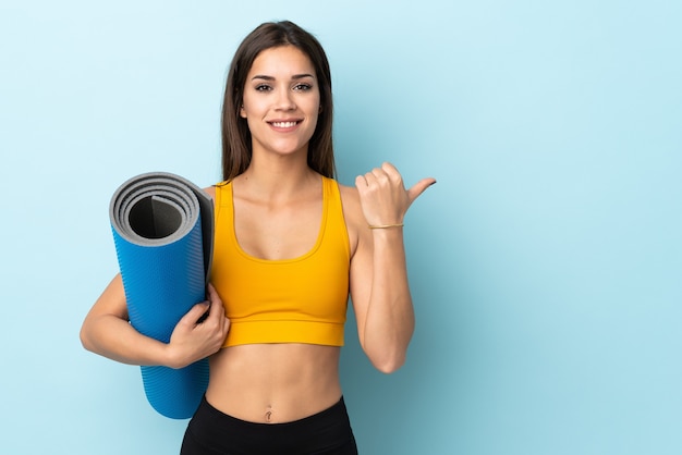 Jeune femme sportive avec tapis isolé sur fond bleu pointant vers le côté pour présenter un produit