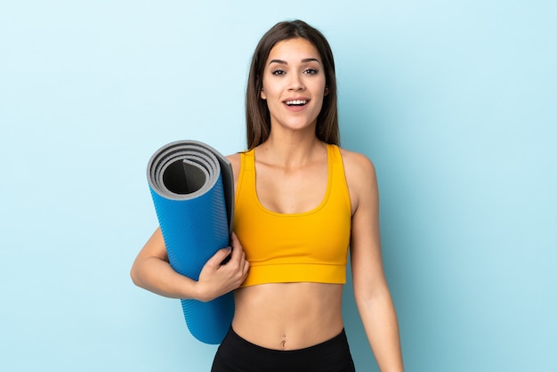 Jeune femme sportive avec tapis isolé sur fond bleu avec surprise et expression faciale choquée