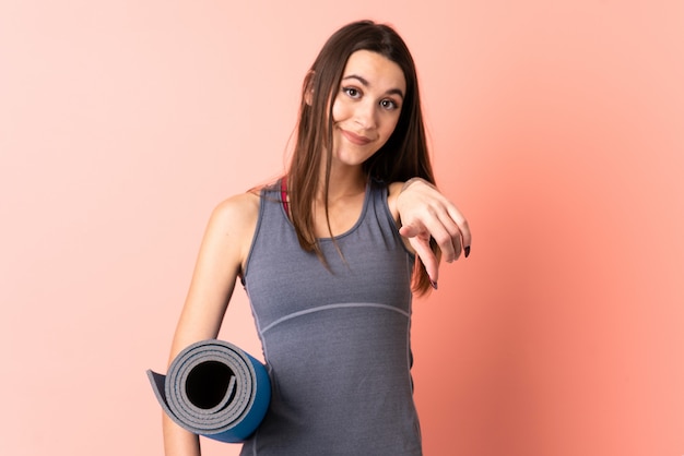 Jeune femme sportive avec tapis sur le mur isolé pointe le doigt vers vous avec une expression confiante