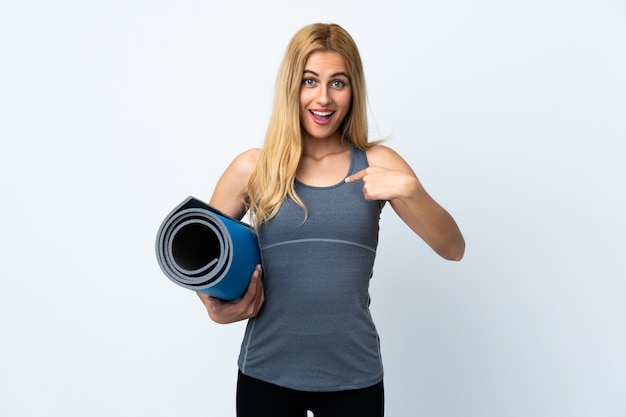 Jeune femme sportive va aux cours de yoga tout en tenant un tapis sur blanc isolé avec expression faciale surprise