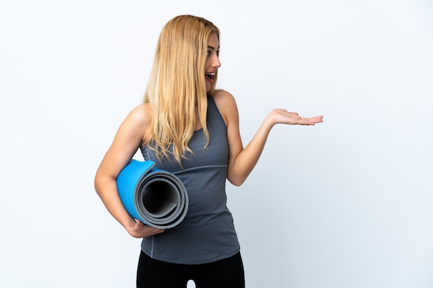 Jeune femme sportive va aux cours de yoga tout en tenant un tapis sur un mur blanc isolé avec une expression faciale surprise