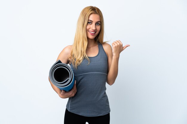 Jeune femme sportive va aux cours de yoga tout en tenant un tapis sur un mur blanc pointant vers le côté