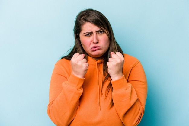 Jeune femme en surpoids caucasienne isolée sur fond bleu montrant le poing à la caméra expression faciale agressive