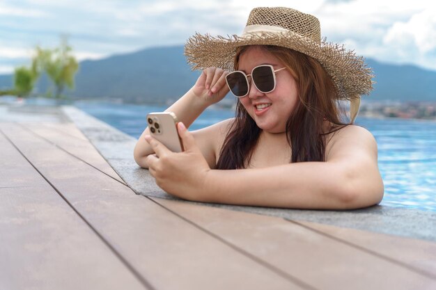Jeune femme en surpoids maillot de bain orange relaxant et tenant un smartphone dans la piscine Femme obèse vacances voyageant main en utilisant le téléphone