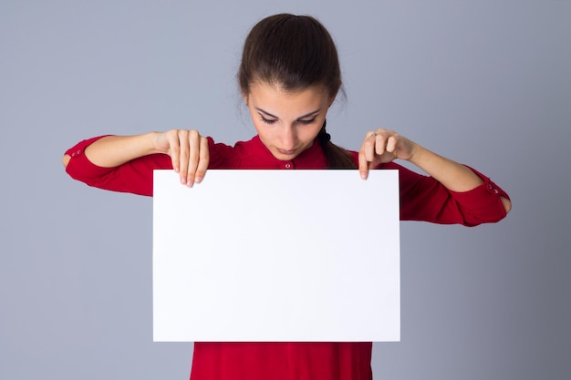 Jeune femme surprise en blouse rouge cachant la moitié de son visage par une feuille de papier blanche en studio