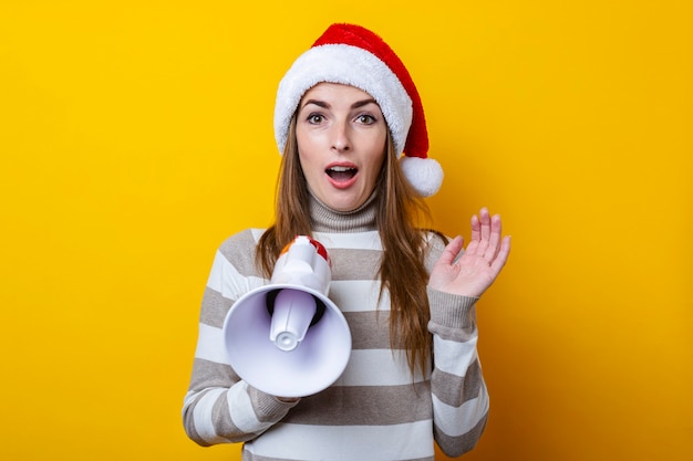 Jeune femme surprise en chapeau de père Noël avec un mégaphone sur fond jaune.