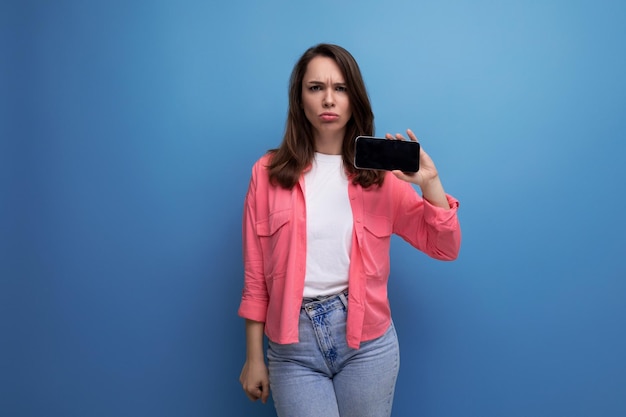 Jeune femme surprise en chemise rose et jeans avec téléphone sur fond isolé de studio