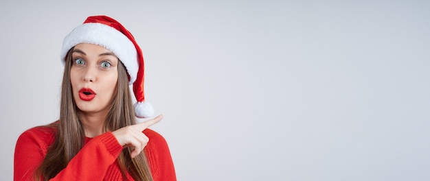 Une jeune femme surprise dans un bonnet de Noel, vêtu d'un pull rouge, pointe son index sur le côté. Bannière avec espace de copie pour votre publicité.