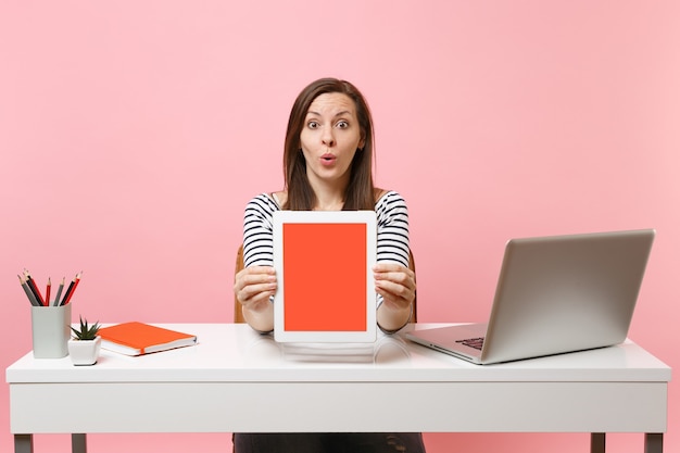 Jeune femme surprise montrant un ordinateur tablette avec un écran vide vierge s'asseoir au bureau blanc avec un ordinateur portable pc contemporain