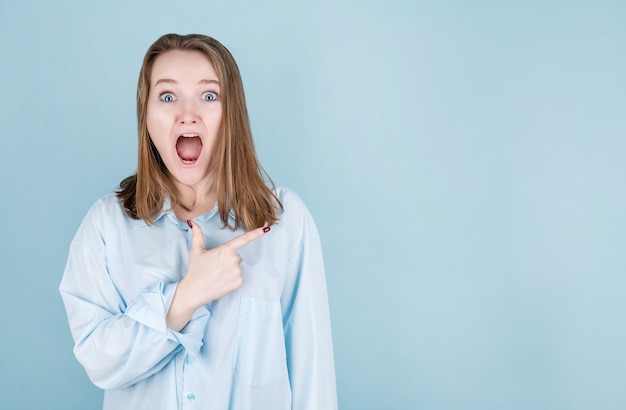 Une jeune femme surprise pointe son doigt vers l'endroit à sa gauche. Portrait d'une fille surprise sur bleu.