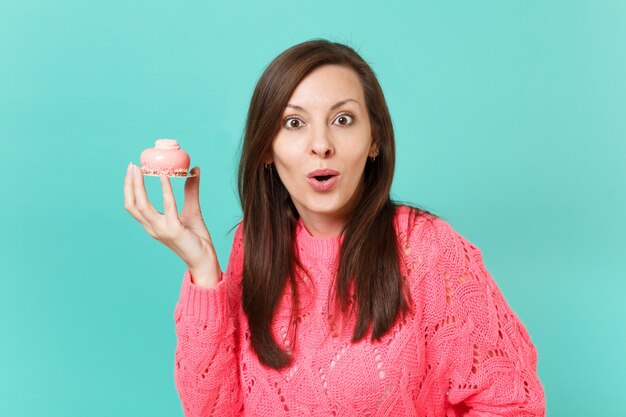 Jeune femme surprise en pull rose tricoté, gardant la bouche grande ouverte, tenant à la main un gâteau isolé sur fond de mur bleu turquoise, portrait en studio. Concept de mode de vie des gens. Maquette de l'espace de copie.