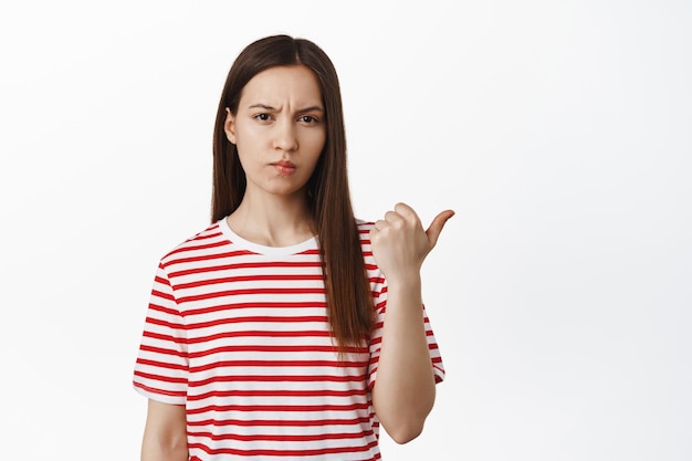 Jeune femme suspecte et sceptique, fronçant les sourcils et plissant les yeux avec incrédulité, pointant le doigt vers qch suspect ou douteux, debout sur fond blanc.