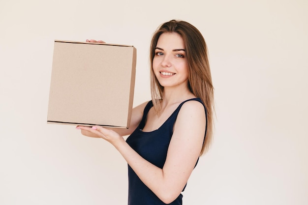 Jeune femme svelte en robe noire sur fond beige avec boîte en carton dans ses mains libre