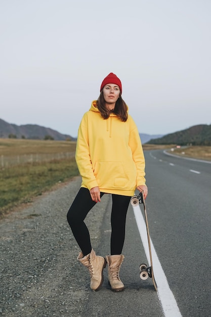 Jeune femme en sweat à capuche jaune et chapeau rouge sur planche à roulettes sur route contre le magnifique paysage de montagne