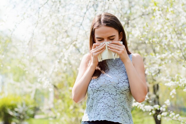 Jeune femme avec des symptômes d'éternuements coronavirus debout dans un verger en fleurs. Jeune, malade, avoir, grippe, courant, nez, fièvre, debout, ressort, Parc