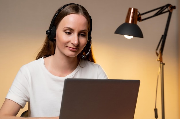 Jeune femme en T-shirt blanc et casque travaille sur un ordinateur portable à la maison Travail à domicile Vidéoconférence Appel vidéo Cours en ligne pour les étudiants