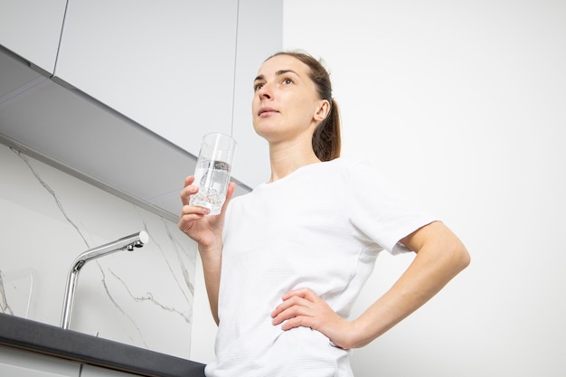 Jeune femme en t-shirt blanc tenant un verre d'eau dans la cuisine