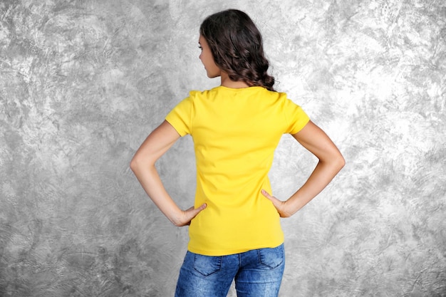 Jeune femme en t-shirt jaune blanc debout contre un mur texturé gris