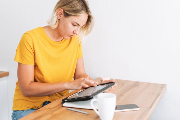 Jeune femme en t-shirt jaune étudie les graphiques de crypto-monnaie et la cotation boursière sur tablette numérique à la maison