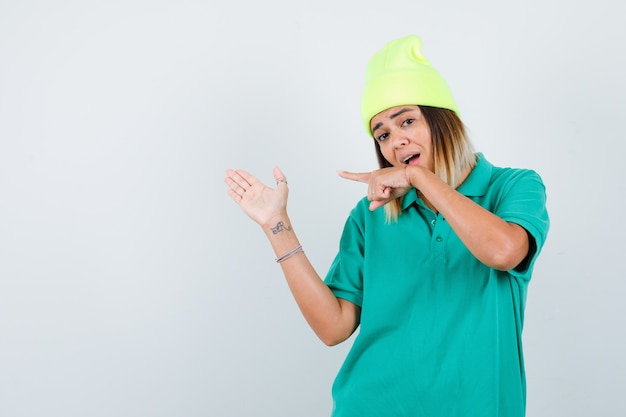 Jeune femme en t-shirt polo, bonnet pointant vers la gauche, montrant un geste de bienvenue et l'air surpris, vue de face.