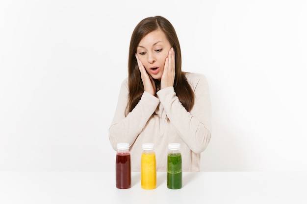 Jeune femme à table avec des smoothies de désintoxication verts, rouges et jaunes dans des bouteilles isolées sur fond blanc. Bonne nutrition, boisson végétarienne, mode de vie sain, concept de régime. Zone avec espace de copie.