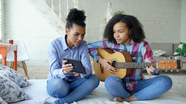 jeune femme avec tablette assise sur le lit enseignant à sa sœur adolescente à jouer de la guitare acoustique