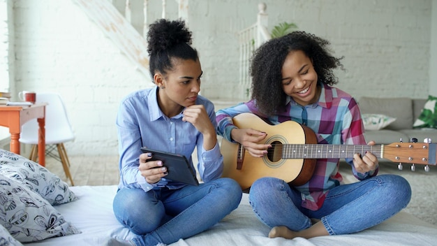 Photo jeune femme avec tablette assise sur le lit enseignant à sa sœur adolescente à jouer de la guitare acoustique