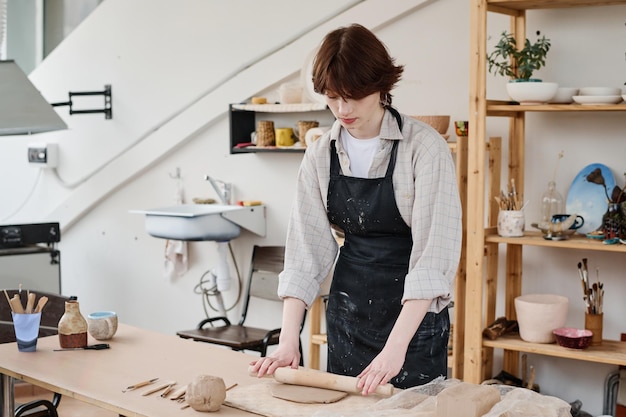 Jeune femme en tablier noir aplatissant le morceau d'argile avec un rouleau à pâtisserie en atelier