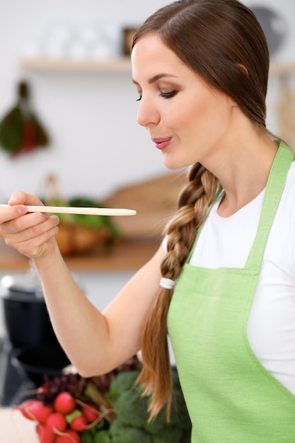 Jeune femme en tablier vert cuisine dans une cuisine La femme au foyer goûte la soupe à la cuillère en bois