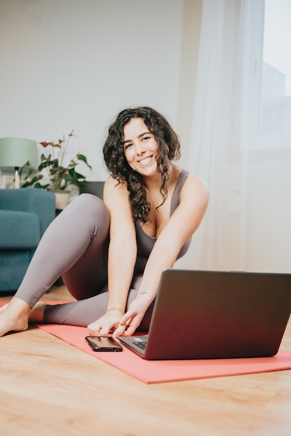 Jeune femme de taille plus assise sur le sol se reposant après avoir fait un cours de yoga en ligne avec son ordinateur portable. Souriant à la caméra et heureux de perdre du poids avec les cours. Vêtements sportifs.