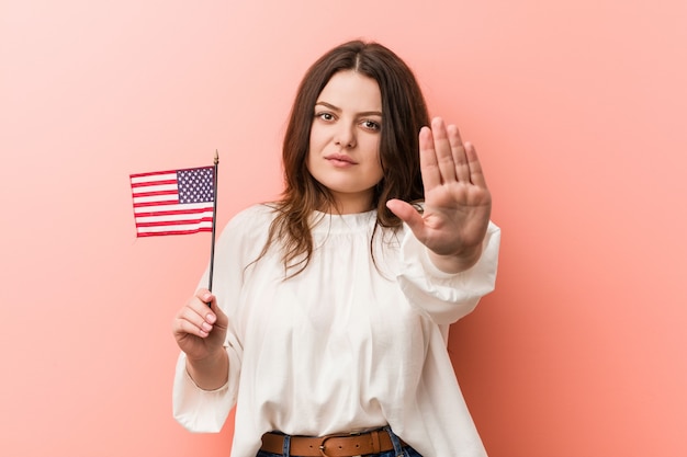 Jeune femme de taille plus curviligne tenant un drapeau des états-unis debout avec la main tendue montrant le panneau d&#39;arrêt, vous empêchant.