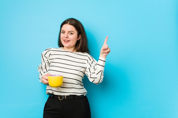 Jeune femme de taille plus tenant une tasse de thé souriant joyeusement pointant avec l'index loin.