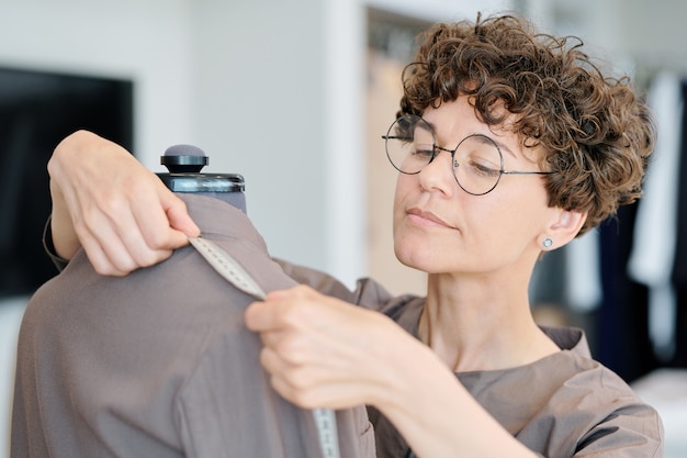 Jeune femme tailleur mesurant la longueur d'épaule de veste inachevée sur mannequin en atelier