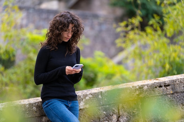 Jeune femme tapant un message sur son portable alors qu'elle est seule dans le parc Elle a les cheveux bruns bouclés porte un pull noir et un jean bleu