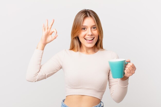 Jeune femme avec une tasse de café se sentant heureuse, montrant son approbation avec un geste correct