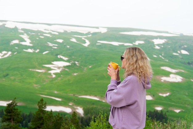 Une jeune femme avec une tasse jaune dans ses mains regarde un plateau dans les montagnes Vacation