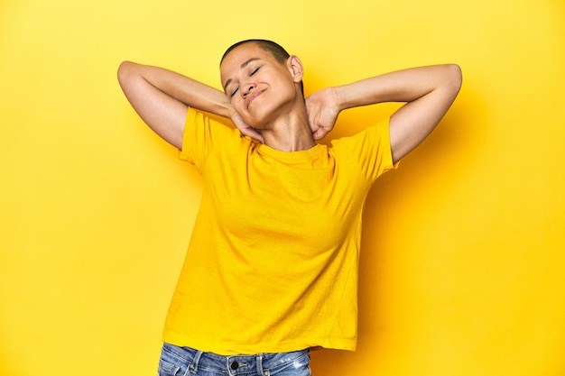 Jeune femme en tee jaune toile de studio jaune se sentant confiante avec les mains derrière la tête