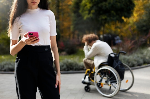 Jeune femme avec un téléphone portable dans ses mains au premier plan sur un arrière-plan flou un gars tenant sa tête dans ses mains assis bouleversé dans un fauteuil roulant