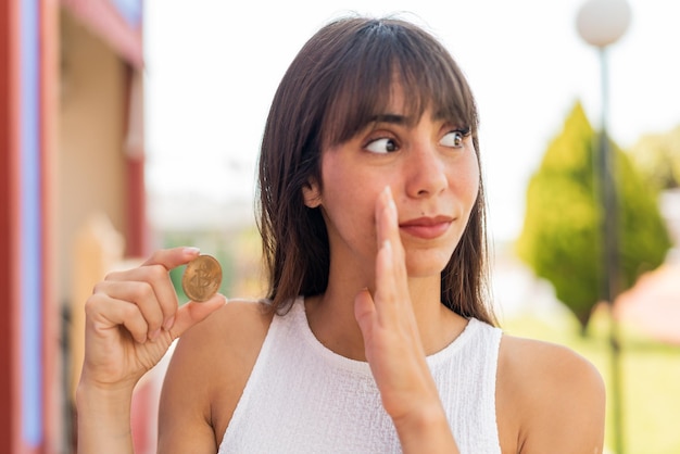 Jeune femme tenant un bitcoin à l'extérieur en chuchotant quelque chose