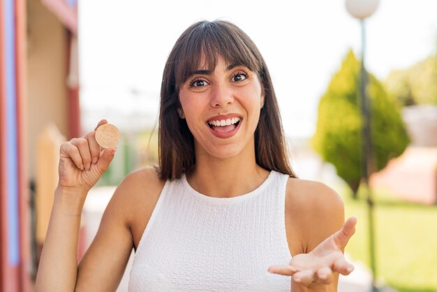 Jeune femme tenant un Bitcoin à l'extérieur avec une expression faciale choquée